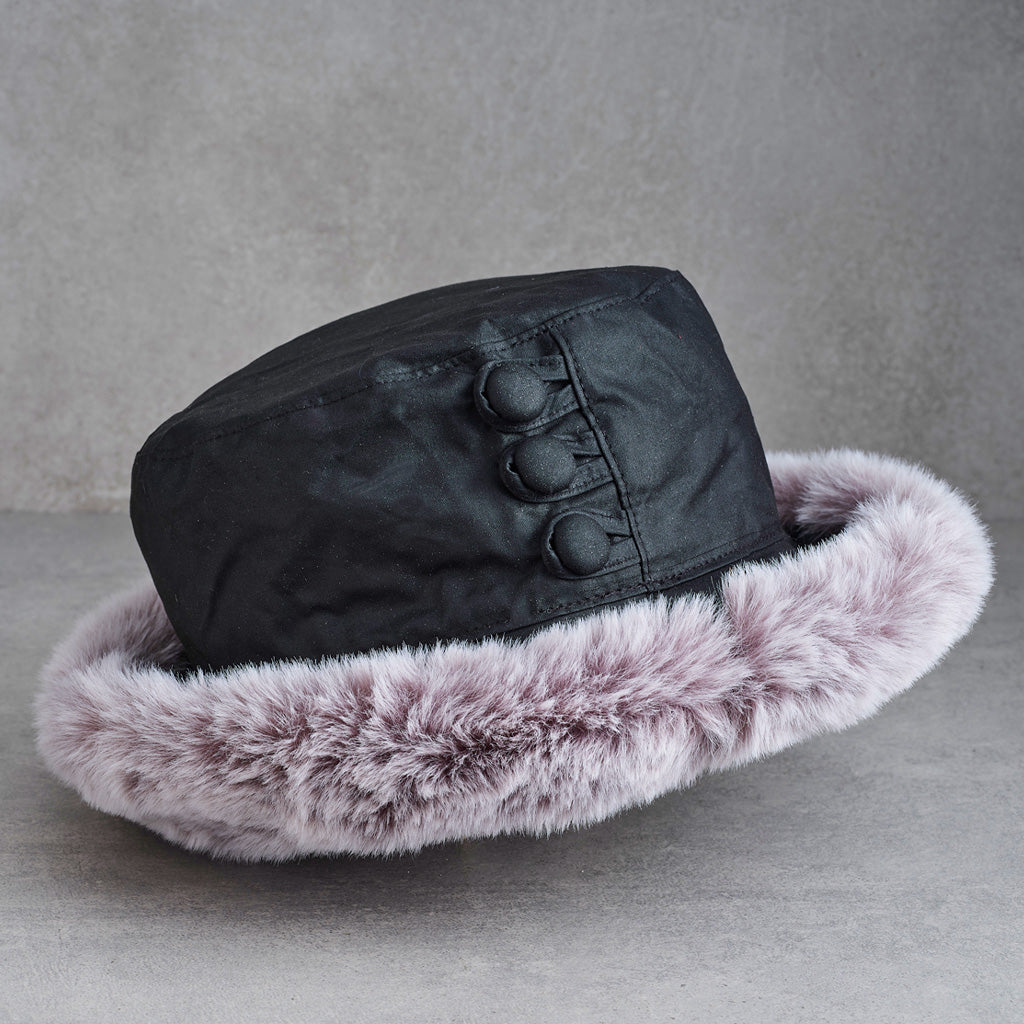 A black winter hat with a plush light pink faux fur trim and three decorative buttons on the side, displayed on a textured gray surface.