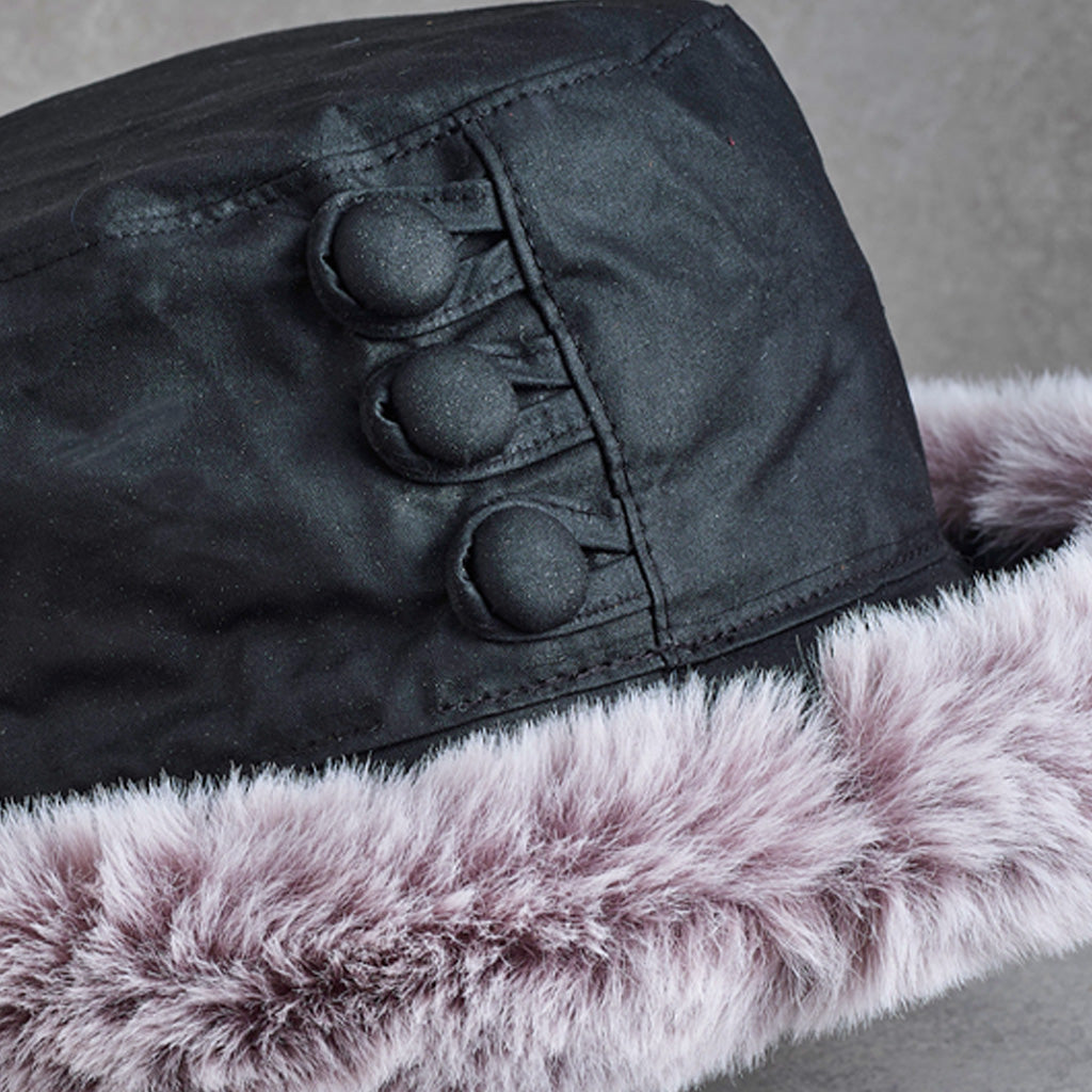 Close-up of a winter hat featuring a black fabric crown with three decorative button details and a thick, fluffy light pink faux fur trim.