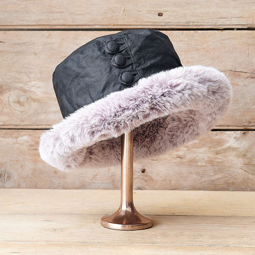 A black fabric hat with a wide light gray faux fur trim, displayed on a metallic stand against a rustic wooden background.