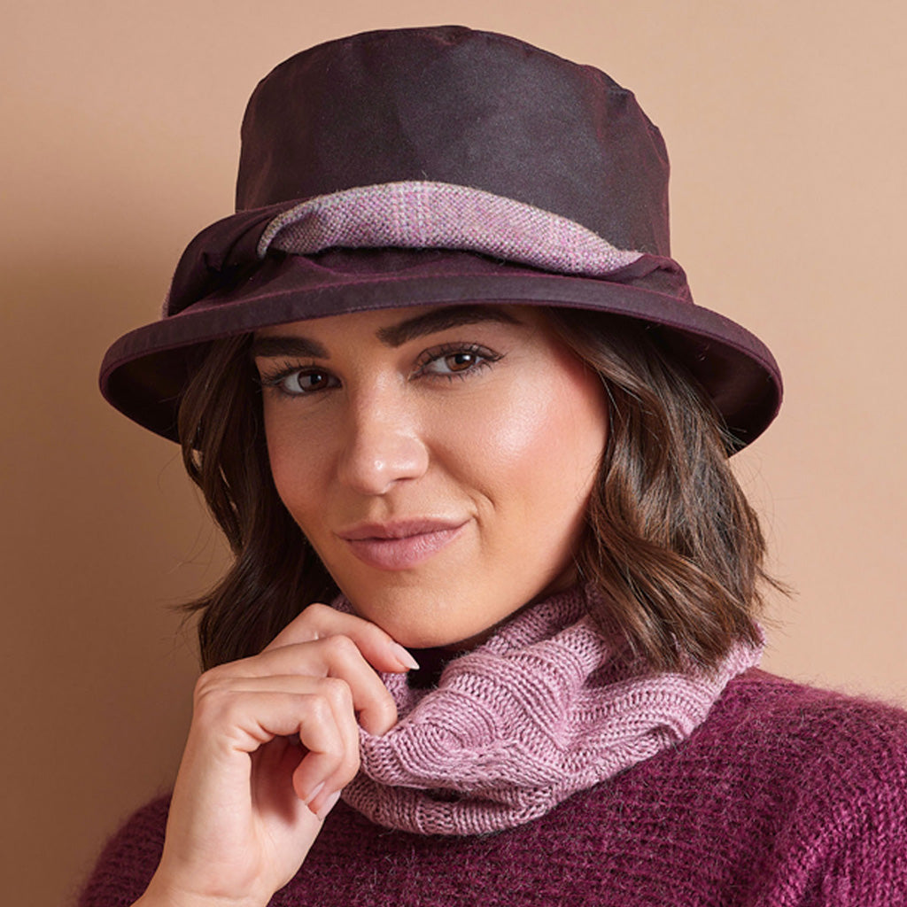 A woman wearing a dark plum-colored wide-brim hat with a textured mauve band, a matching mauve knit cowl neck warmer, and a plum knit sweater, set against a solid warm beige background.