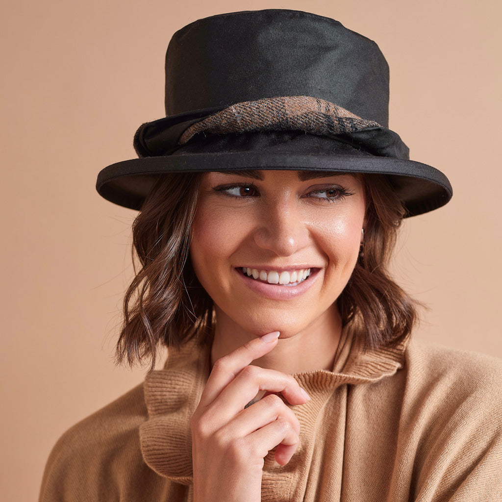 A smiling woman wearing a black wide-brim hat with a textured brown band and a camel-colored knit top, set against a solid beige background.