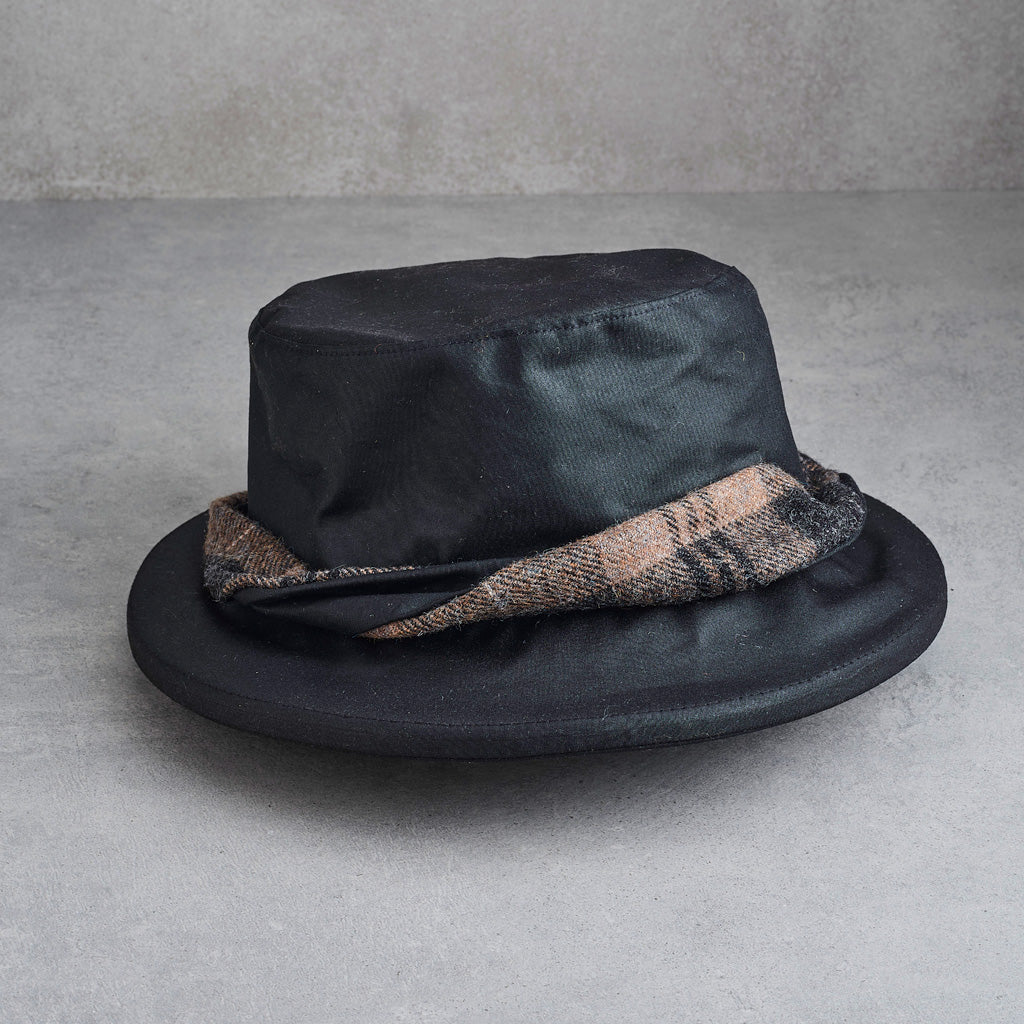 A black fedora hat with a textured plaid band in beige, brown, and black, resting on a gray concrete surface.
