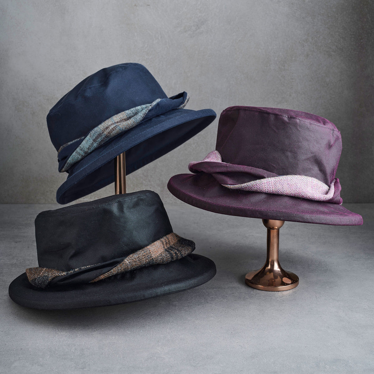 Three wide-brim fedora hats displayed on metallic stands against a textured gray studio background. One hat is dark navy blue, one is black, and one is muted purple, each with a patterned fabric band.