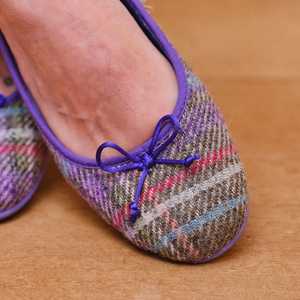 A close-up of a person's foot wearing a purple-trimmed flat shoe with a multicolor plaid pattern and a matching purple bow on the toe, set against a warm wooden floor.