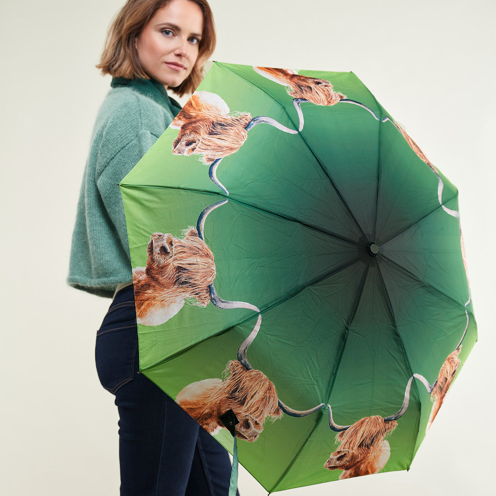 A woman holding a green umbrella with a repeating print of Highland cattle, photographed against a plain light background.