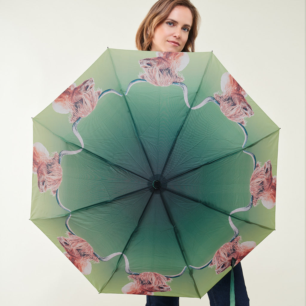 A woman holds up a large open umbrella showing its underside, which features a green gradient design with a repeating pattern of pink and brown animal faces.