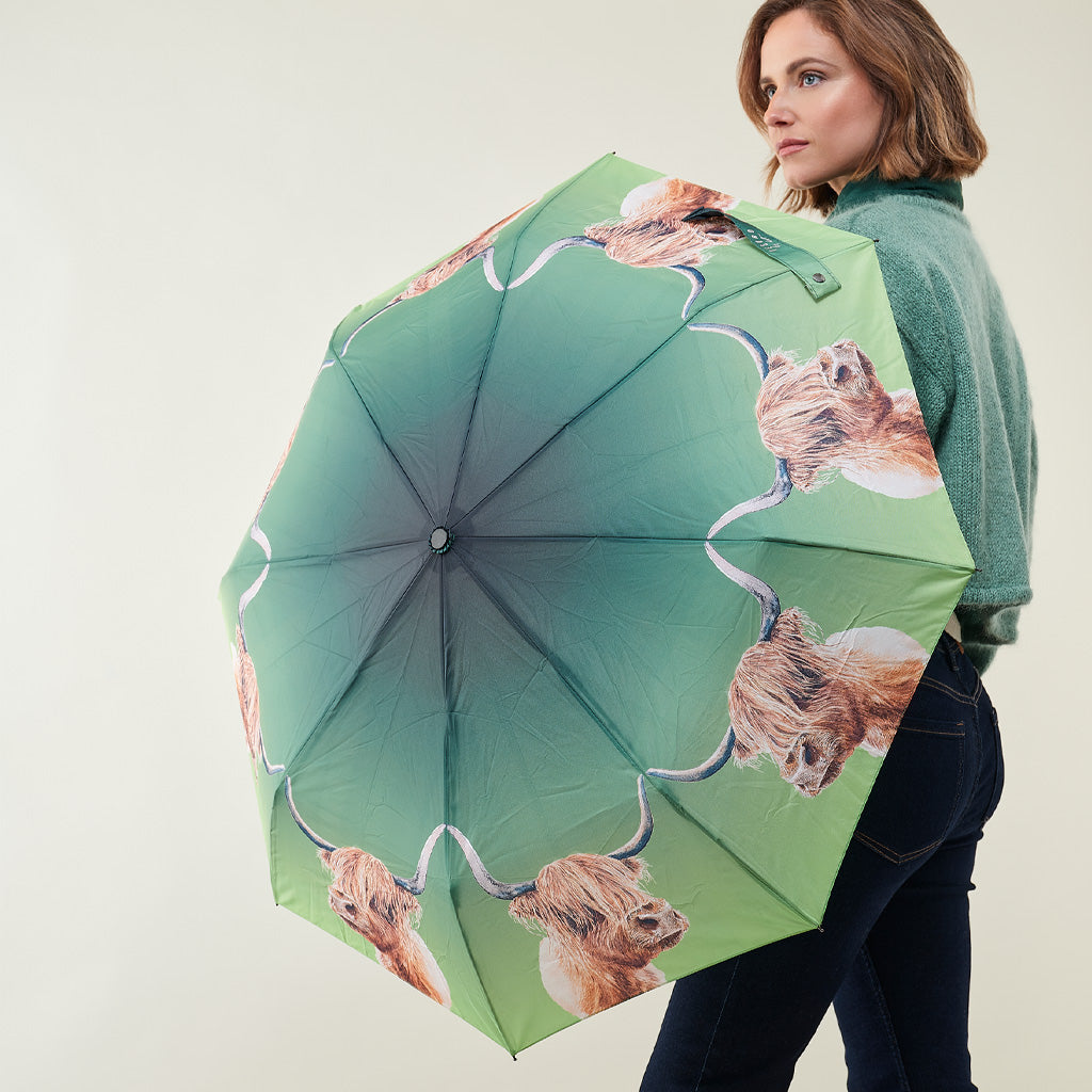 A woman holding a large open umbrella with a green gradient background and a repeating Highland cow print. The woman is wearing a green sweater and dark jeans, photographed against a plain light gray studio background.