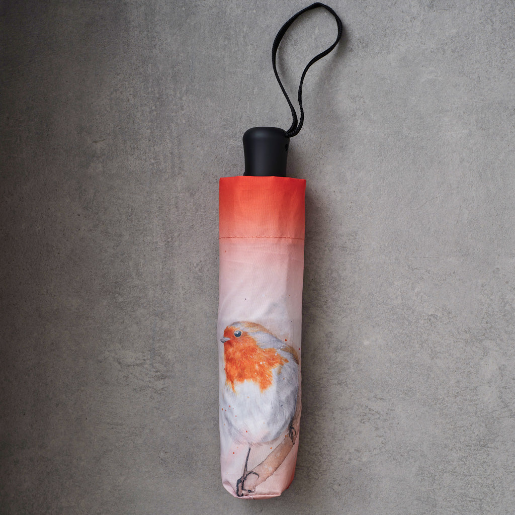 A folded compact umbrella with a black handle and wrist strap, featuring a printed design of a white bird with orange and pink tones, resting on a textured gray concrete surface.