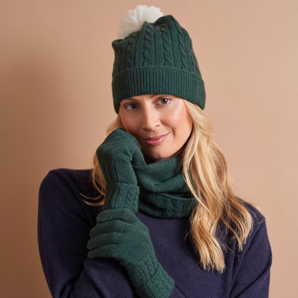 A woman wearing a dark green cable-knit beanie with a white pom-pom, a matching dark green knit scarf, and dark green knit gloves, posed against a solid warm beige background.