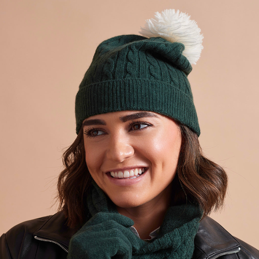 A woman wearing a dark green cable-knit beanie with a white pom-pom, a matching dark green knit scarf, and dark green knit gloves, posed against a solid warm beige background.