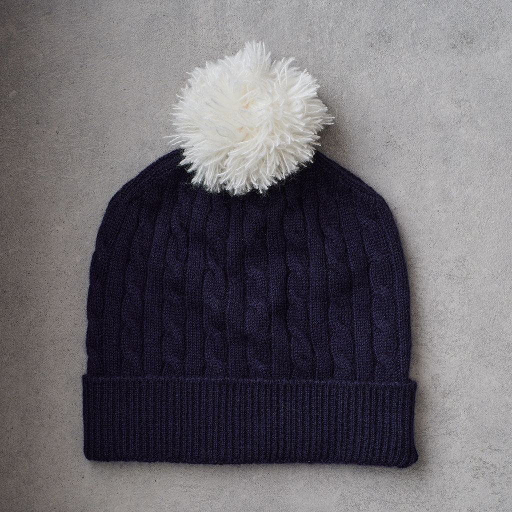 A navy blue cable-knit beanie hat with a ribbed cuff and a fluffy white pom-pom on top, laid flat on a textured gray surface.