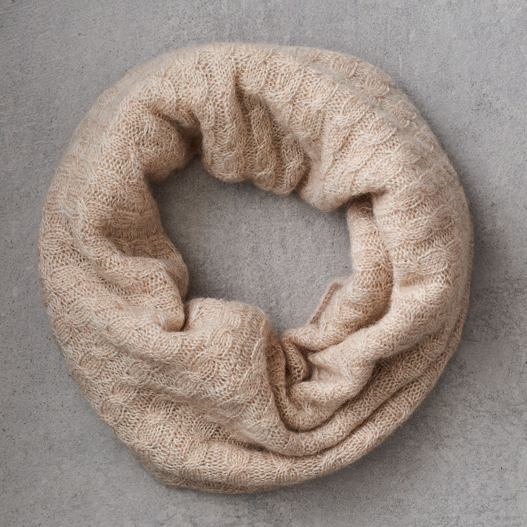 A beige knit neckwarmer with a textured cable pattern, arranged in a circle on a neutral gray background.