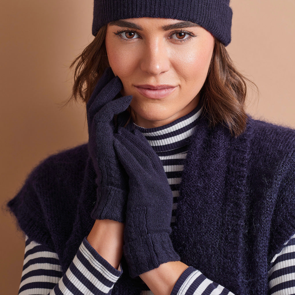 A woman wearing a dark navy knit beanie, matching knit gloves, a navy sleeveless sweater vest, and a black and white striped turtleneck sweater, posed against a solid warm beige background.