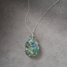 A silver chain necklace with a teardrop-shaped pendant made of iridescent abalone shell, displayed on a textured gray background.
