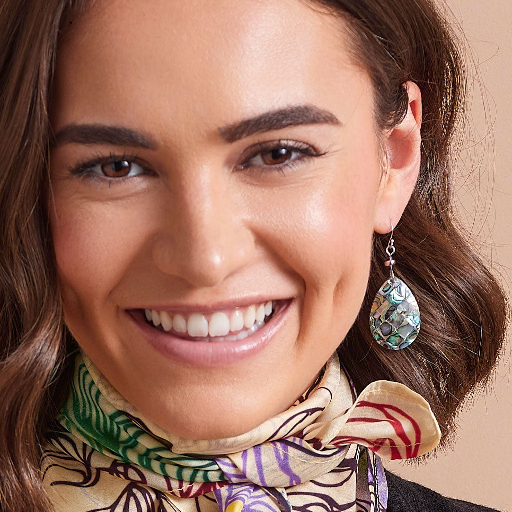 A close-up of a smiling woman wearing a dangling teardrop earring with a green, blue, and white patterned stone setting, along with a patterned silk scarf.