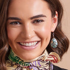 A close-up of a smiling woman wearing a dangling teardrop earring with a green, blue, and white patterned stone setting, along with a patterned silk scarf.
