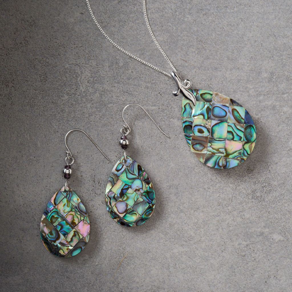 A jewelry set featuring a silver necklace and matching drop earrings, both with teardrop-shaped pendants made of iridescent abalone shell mosaic. The jewelry is displayed on a textured gray background.