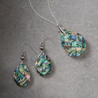 A jewelry set featuring a silver necklace and matching drop earrings, both with teardrop-shaped pendants made of iridescent abalone shell mosaic. The jewelry is displayed on a textured gray background.