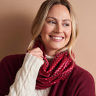 A smiling blonde woman wearing a red patterned infinity scarf and a burgundy knit sweater over a cream cable-knit top, posed against a solid warm beige background.