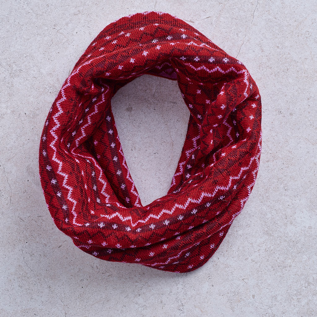 A red infinity scarf with white and dark red geometric patterns, laid flat on a light gray textured background.