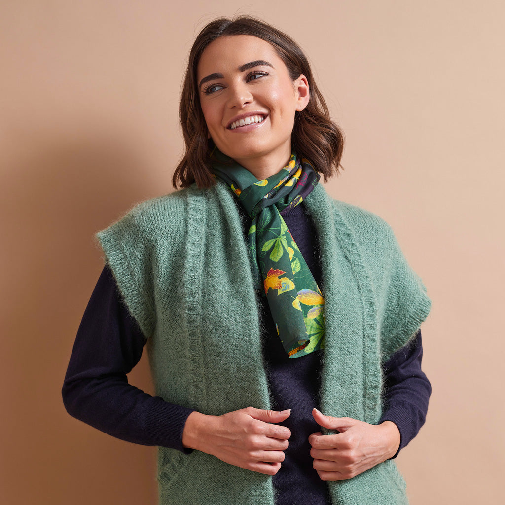 A woman wearing a sage green knit sleeveless vest over a dark navy sweater, accessorized with a dark green scarf featuring a colorful animal print, set against a solid beige background.