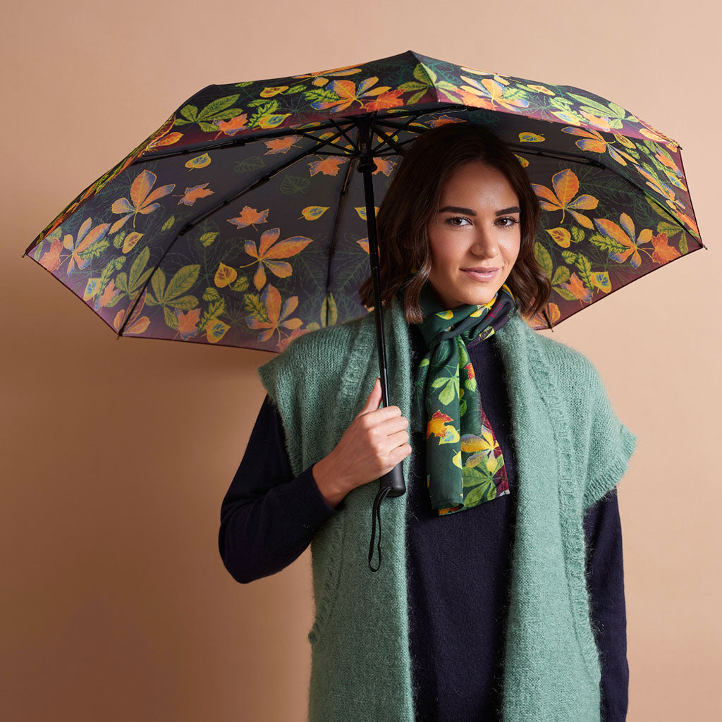 A woman holding a black umbrella with a vibrant orange and green floral pattern, styled with a green knit vest and matching scarf against a solid beige background.