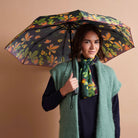 A woman holding a black umbrella with a vibrant orange and green floral pattern, styled with a green knit vest and matching scarf against a solid beige background.