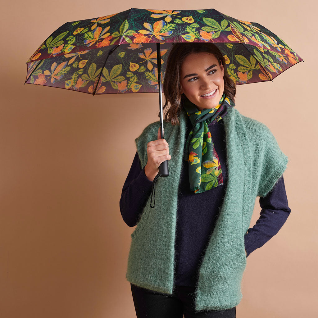 A woman holding a dark umbrella with a colorful orange and green leaf pattern, wearing a fuzzy sage green sleeveless vest over a navy sweater and a matching patterned scarf, set against a solid tan studio background.