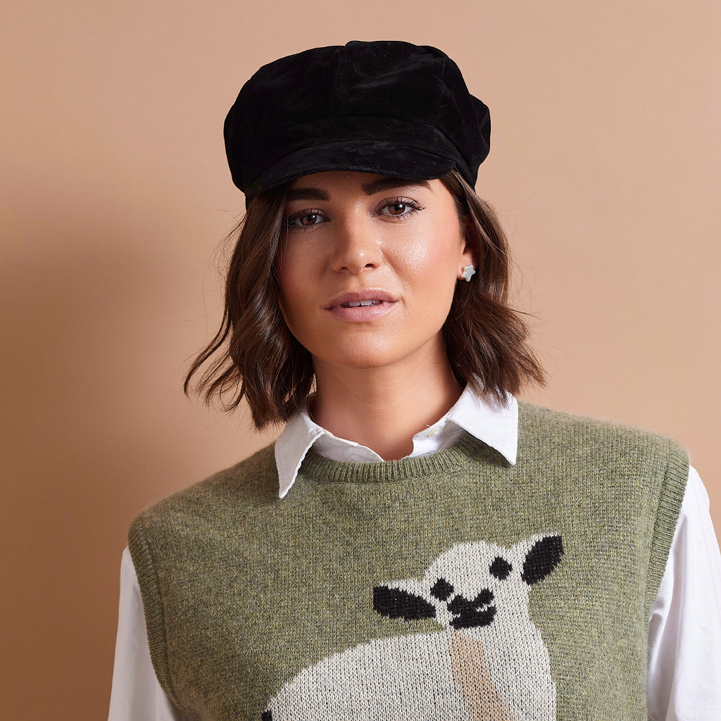 A woman wearing a black velvet newsboy cap, an olive green knit sweater vest with a white and black sheep motif, and a white collared shirt, set against a solid warm beige background.