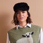 A woman wearing a black velvet newsboy cap, an olive green knit sweater vest with a white and black sheep motif, and a white collared shirt, set against a solid warm beige background.