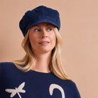 A woman wearing a navy blue newsboy cap and a matching navy blue knit sweater with white graphic details, set against a solid beige background.