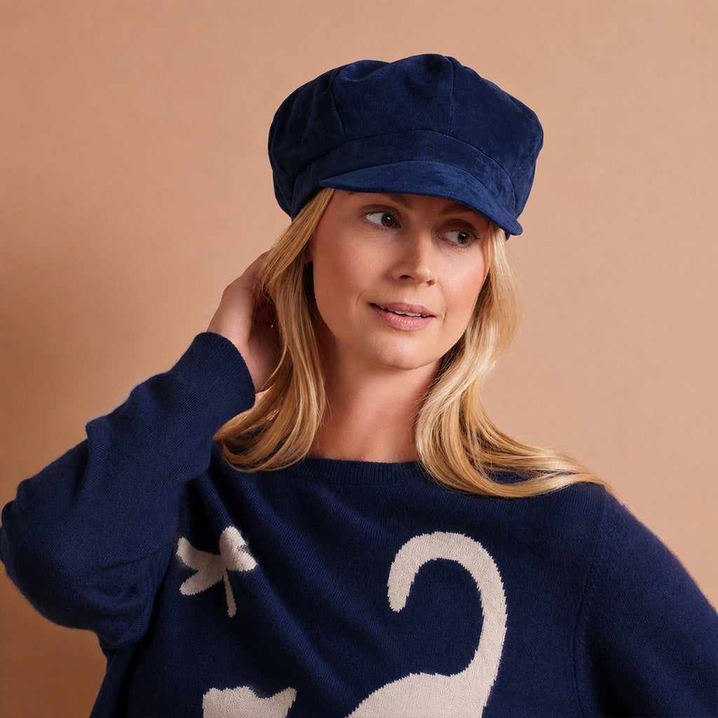 A woman wearing a navy blue knit sweater with a white cat and dragonfly motif, paired with a matching navy blue velvet newsboy cap, set against a solid warm beige background.
