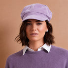 A woman wearing a lavender purple newsboy cap, a matching purple knit sweater, and a white collared shirt, set against a solid warm tan background.