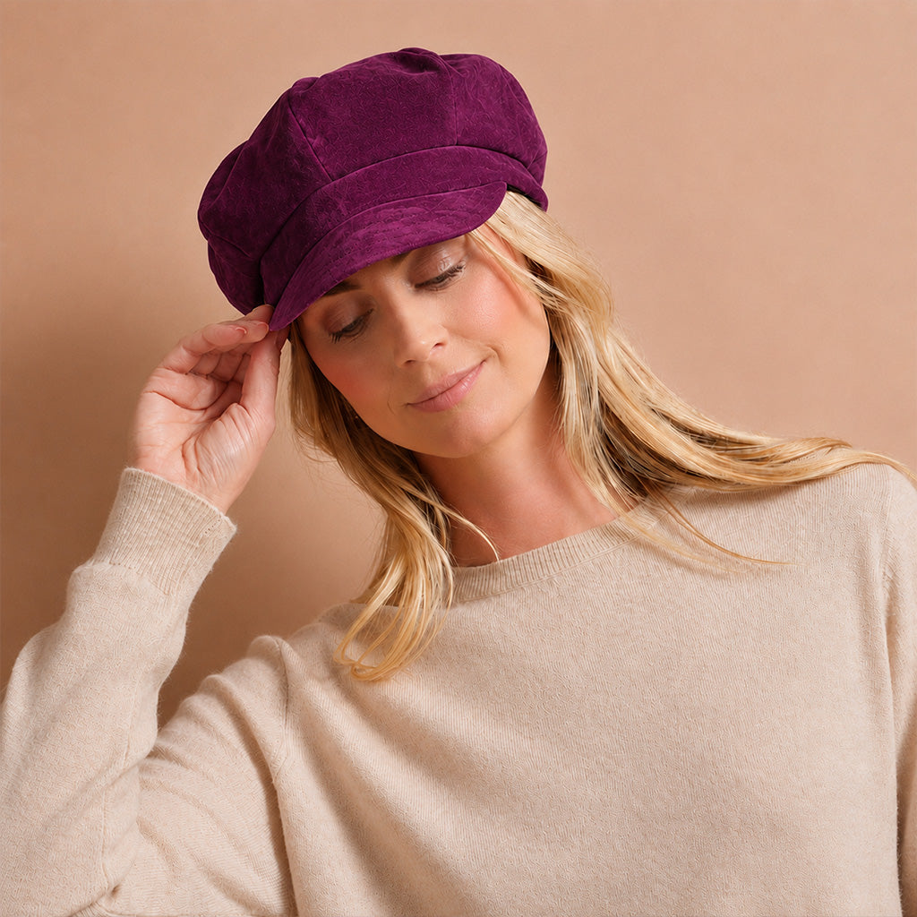 A woman wearing a deep purple velvet newsboy cap and a beige knit sweater, posing against a solid warm beige background.