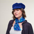 A woman wearing a royal blue velvet newsboy cap and a blue and purple patterned silk scarf, styled with a dark cardigan over a white top, against a plain light beige background.