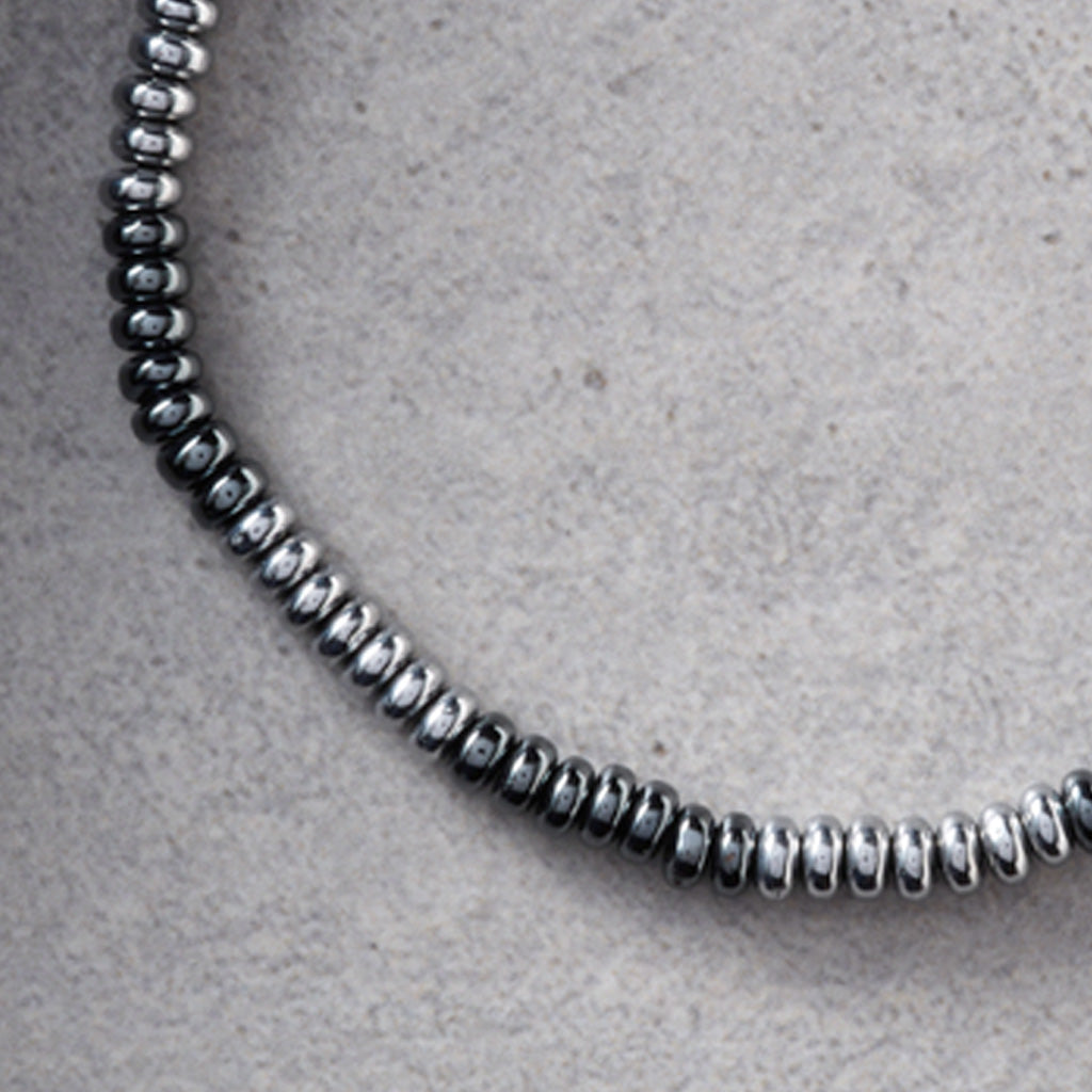 A close-up of a silver-toned beaded chain necklace, showing the metallic cylindrical beads arranged in a curve on a textured light gray surface.