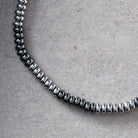 A close-up of a silver-toned beaded chain necklace, showing the metallic cylindrical beads arranged in a curve on a textured light gray surface.