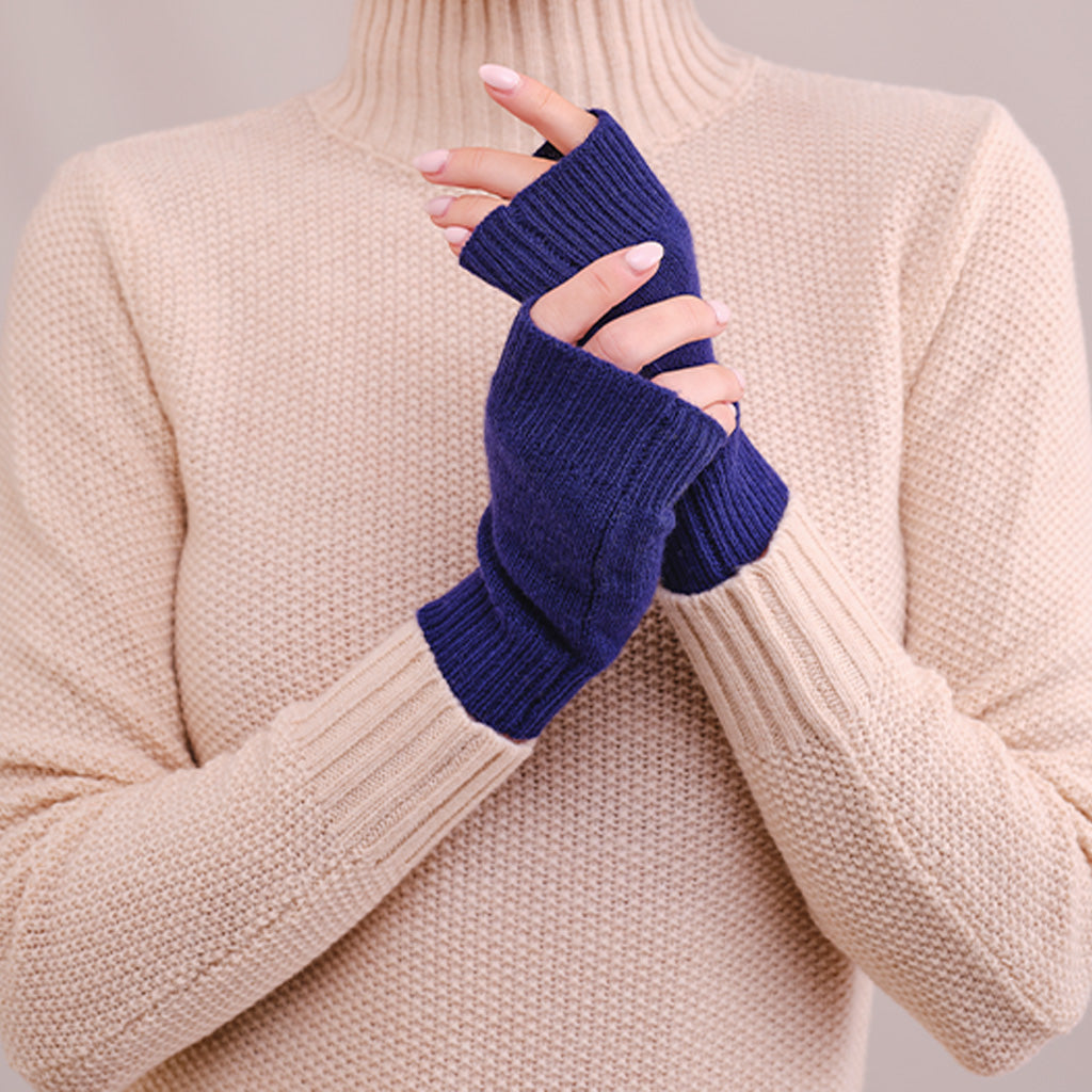 A person wearing a beige ribbed knit turtleneck sweater and dark navy blue fingerless knit gloves, holding their hands together against a neutral light gray background.
