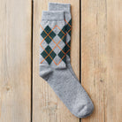 A single gray knit sock with an argyle pattern in dark green and orange, laid flat on a light wood plank background.
