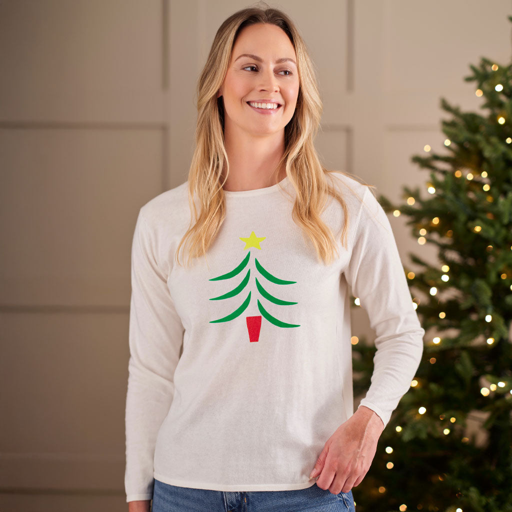 A smiling blonde woman wearing a white long-sleeve crewneck shirt with a green Christmas tree graphic and a yellow star, standing in front of a softly lit holiday tree.