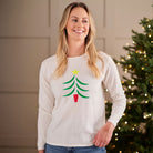 A smiling blonde woman wearing a white long-sleeve crewneck shirt with a green Christmas tree graphic and a yellow star, standing in front of a softly lit holiday tree.