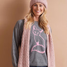 A smiling woman wearing a pink knit beanie, a gray sweater with a pink fox graphic, and a chunky pink cable-knit cardigan, posed against a solid warm beige background.