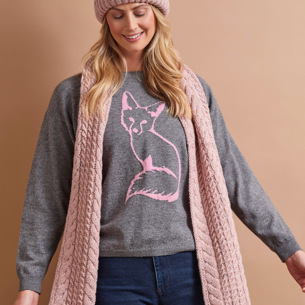 A woman wearing a gray knit sweater with a pink fox graphic, a chunky pink cable-knit cardigan, a pink beanie, and blue jeans, posed against a solid warm beige background.