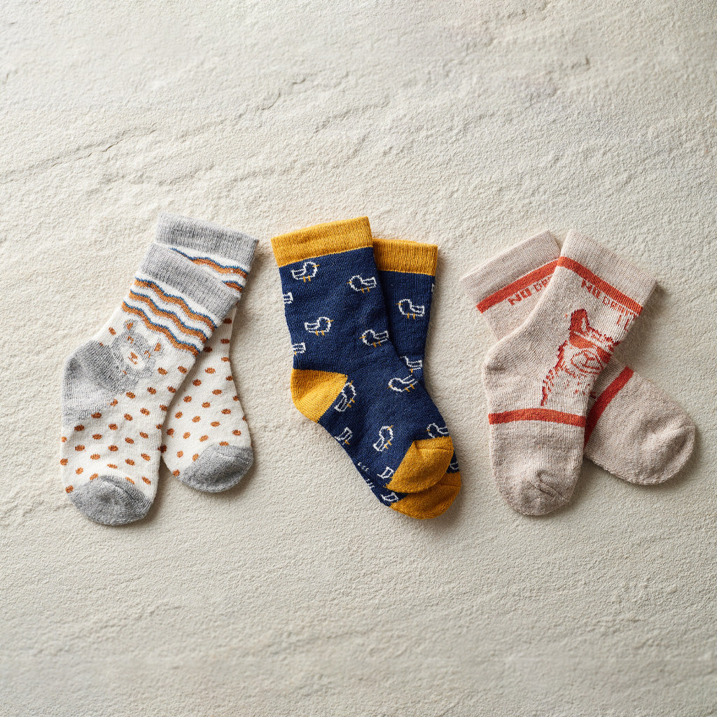 Three pairs of patterned crew socks laid flat on a textured off-white surface: one pair is light gray and white with orange stripes and dots, one pair is navy blue with small white motifs and mustard yellow accents, and one pair is cream with orange stripes and a red graphic.