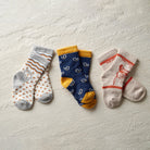 Three pairs of patterned crew socks laid flat on a textured off-white surface: one pair is light gray and white with orange stripes and dots, one pair is navy blue with small white motifs and mustard yellow accents, and one pair is cream with orange stripes and a red graphic.