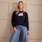 A woman wearing a navy blue crewneck sweatshirt with a white bunny graphic on the chest, paired with wide-leg blue jeans, standing against a beige paneled wall.