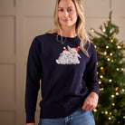 A woman wearing a navy blue crewneck sweater with a white and gray rabbit graphic wearing red Santa hats, standing in front of a softly lit Christmas tree.