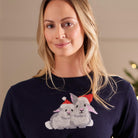 A woman wearing a navy blue crewneck sweater with a knitted graphic of two gray rabbits wearing red Santa hats, set against a softly blurred indoor background.