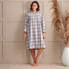 A woman stands barefoot in front of a beige paneled wall wearing a long-sleeve plaid nightshirt with a shawl collar. The nightshirt has a white base with gray and black plaid lines.