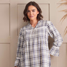 A woman wearing a long-sleeve plaid tunic top with a button-up front and ruffled collar, standing against a beige paneled wall.